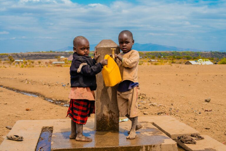 Two children filling a container at a rural water tap on a sunny day.