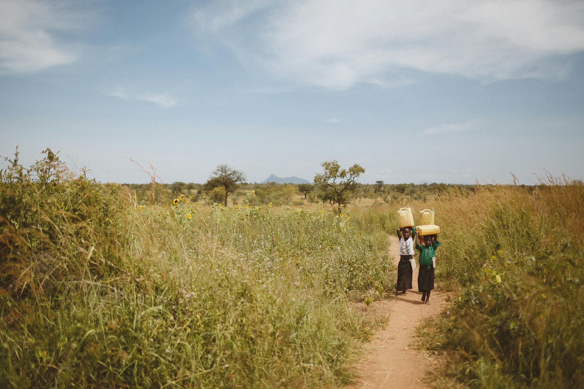 Two children walk through a lush field in Kitgum, Uganda, carrying baskets under a clear sky.